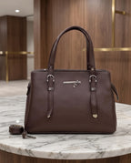 A dark brown leather handbag with silver-tone hardware, displayed on a white marble table with a blurred wooden interior background.