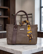 A structured taupe-brown leather tote bag with dark brown handles and trim, featuring a yellow and brown patterned silk scarf tied around the handle, displayed on a white marble countertop.