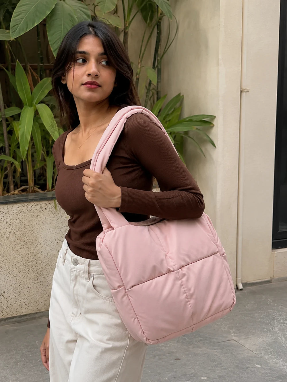 Woman holding a pink puffer bag outdoors with plants and a building in the background