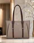 A structured tote bag in taupe with dark brown leather handles and trim, featuring silver-tone hardware and a small rectangular metal plaque on the front, displayed on a marble surface in an elegant indoor setting.