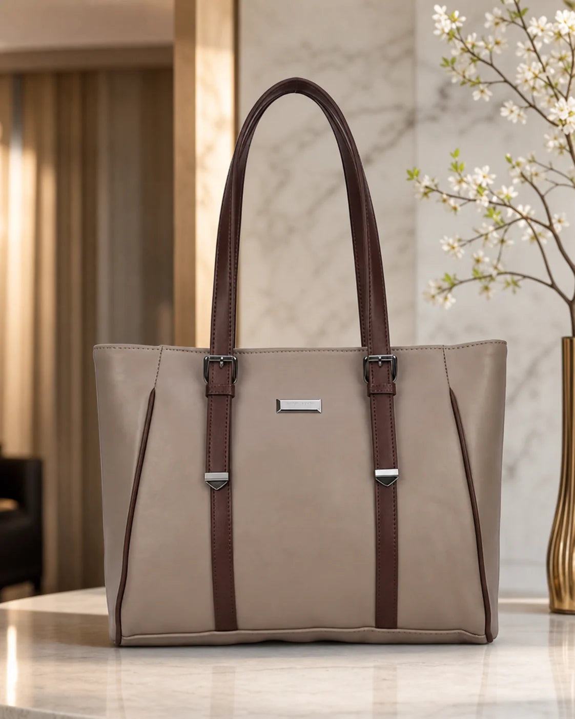 A structured tote bag in taupe with dark brown leather handles and trim, featuring silver-tone hardware and a small rectangular metal plaque on the front, displayed on a marble surface in an elegant indoor setting.