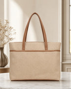 A structured beige tote bag with a textured woven body and smooth tan handles, displayed on a marble surface in a softly lit interior setting.