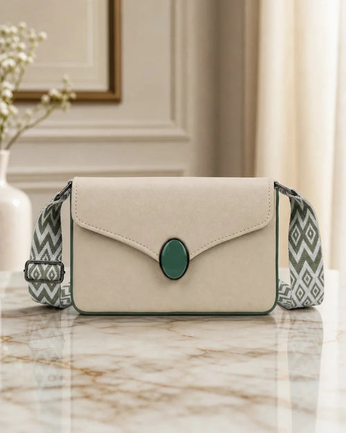 A cream-colored envelope-style crossbody handbag with a green oval center accent and a patterned green and white shoulder strap, resting on a marble surface.